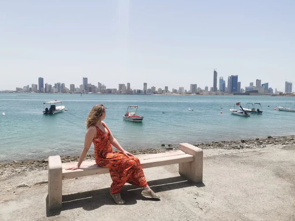 Blonde woman in a long orange sundress sat by the ocean overlooking the Bahrain skyline