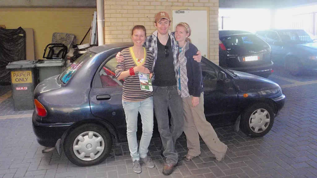 Three people standing outside a small blue hatchback car, ready to go on a roadtrip