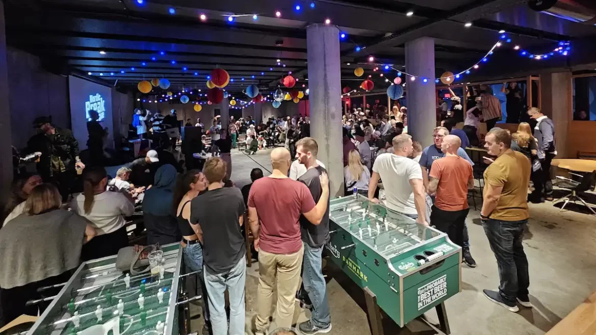 Dozens of people standing in a communal area in a hostel in Copenhagen. There are fairy lights, a large TV and table football.