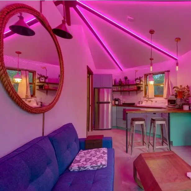 A modern looking open plan living room and kitchen area with a blue sofa, breakfast bar, large mirror on the wall and all bathed in a neon pink light