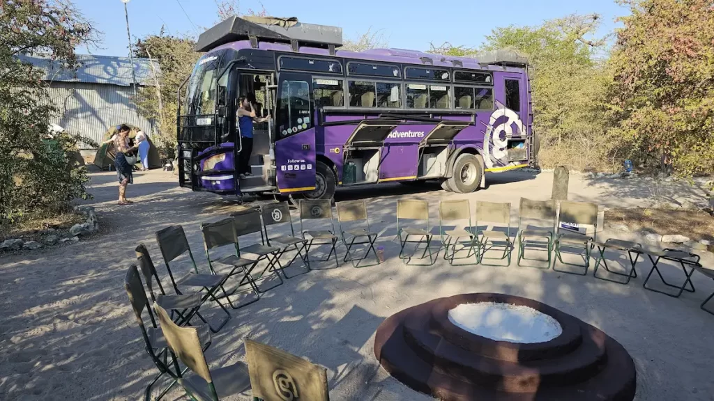 A purple G Adventures tour bus and a circle of camping chairs set up in front of it around an unlit fire pit