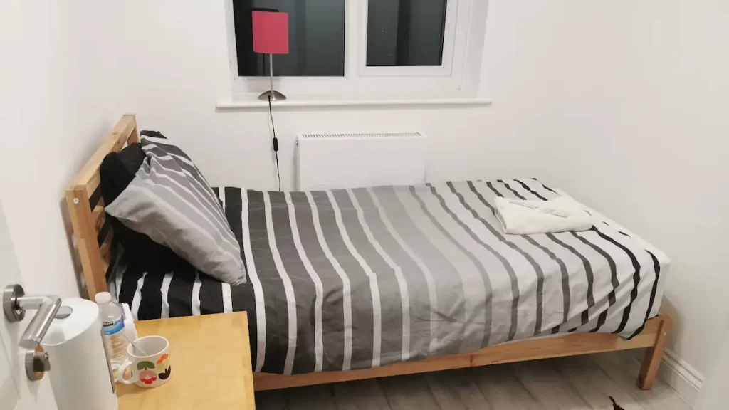 A tiny room with a single bed, a kettle and a lamp. A folded towel sits on the bed