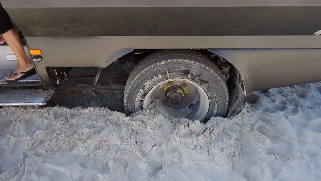 Close up of a tyre wheel showing a van stuck in sand on a beach