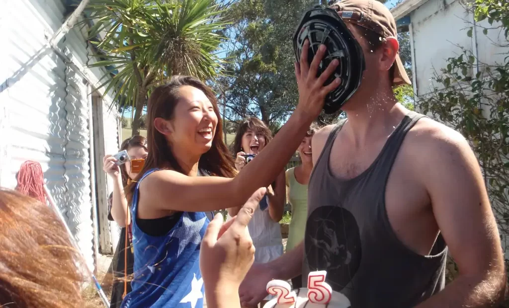 A man getting a cake thrown in his face by a young Japanese girl while holding a birthday cake with candles displaying the number 25. Several women are stood in the background laughing and taking photos