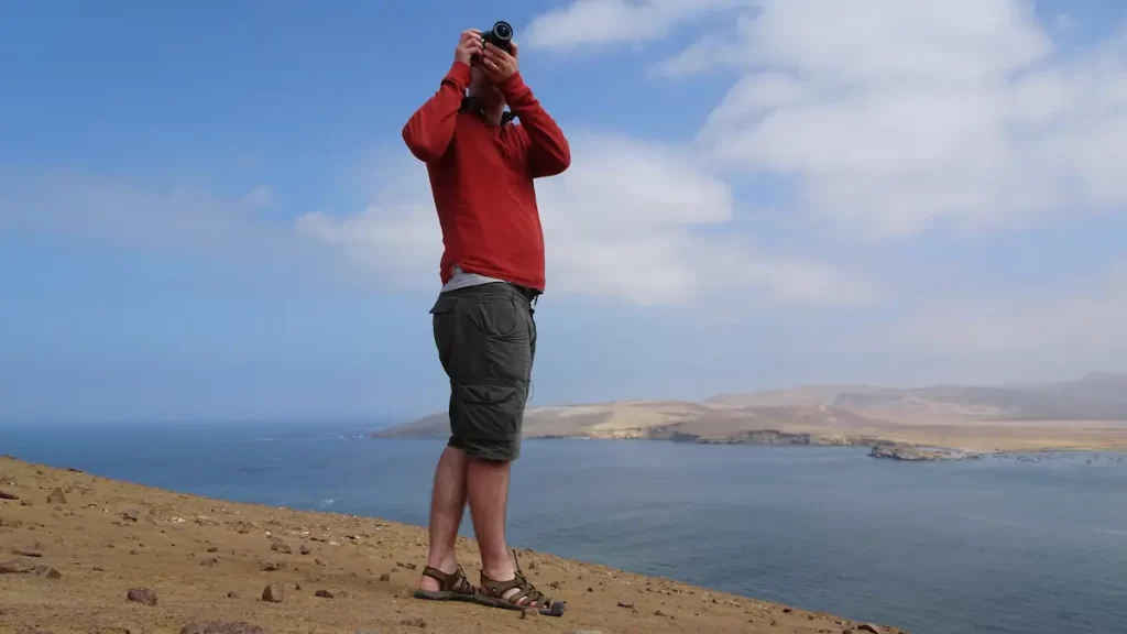Man in a red jumper using a DSLR camera stood on a dusty cliff over looking the ocean