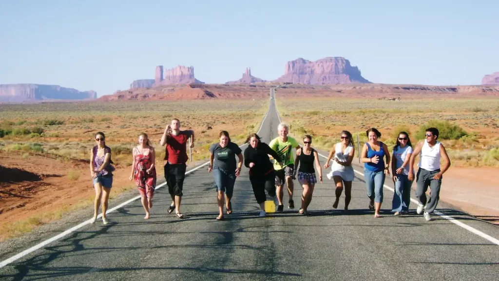 Group of young adults in a line running down a tarmac road with the rock formations of Monument Valley in the background