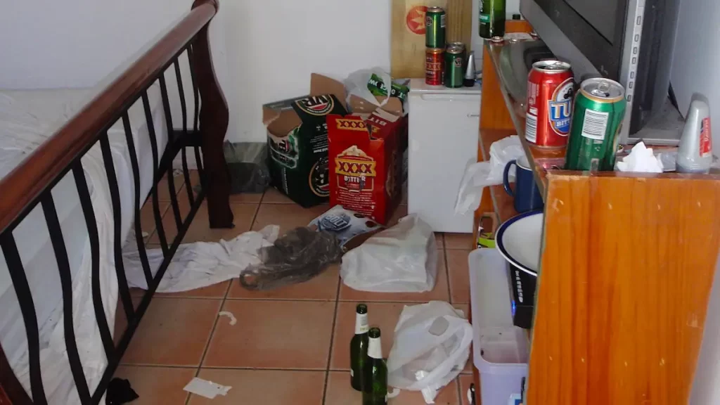 Dirty hostel room with empty beer bottles and beer cans and rubbish scattered all over the floor and surfaces