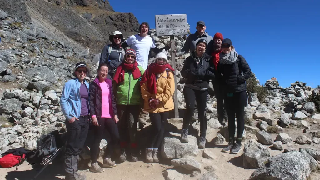 Group of 10 hikers both male and female dressed in brightly coloured warm clothing stood on a mountain in front of a sign that reads 'Abra Salkantay Altitude 4600m'