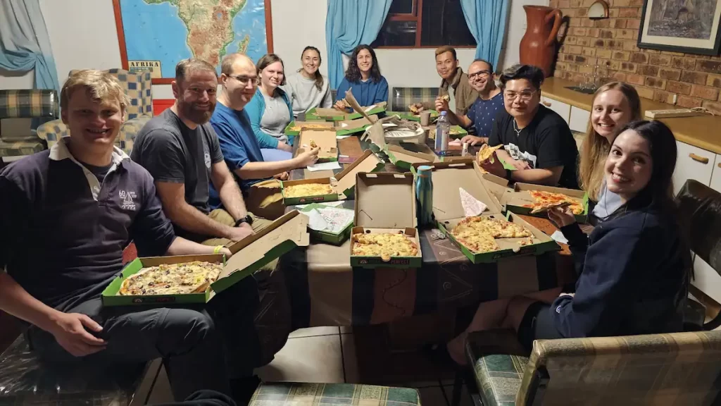 A group of young adults smiling and sat around a table indoors each holding an open pizza box with pizza inside