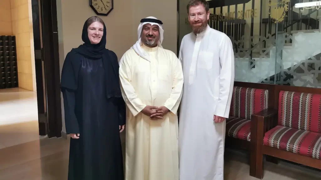 Two men wearing light coloured thobes and a woman wearing a black abaya and hijab standing inside a mosque
