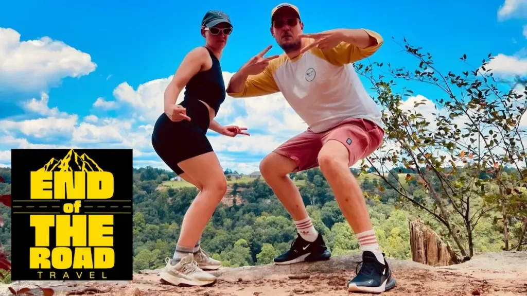 A woman and a man in their mid thirties doing a silly pose and doing the peace sign with their hands. They are stood on a rock with a forest in the background and a blue sky. There is a black and yellow logo on the left hand side of the image which reads 'end of the road travel'.