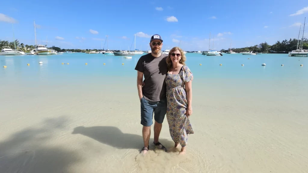Couple stood together at the beach with their feet in the water. The sand is golden and the water is crystal clear and turquiose. There are sailboats and yachts in the background.