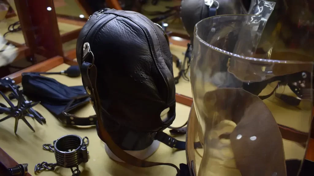 A collection of sex parafernalia in a museum cabinet including a black leather mask, chains and a metal spiked tool