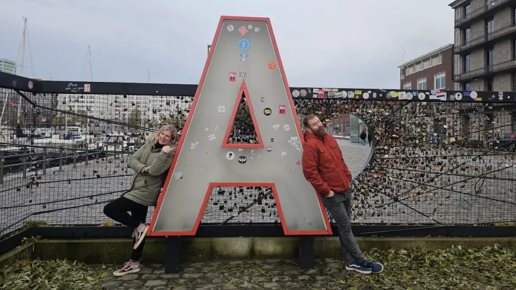 A giant 'A' sign with a woman standing to the left of it and a man standing to the right. Behind them is a black metal fence covered in padlocks