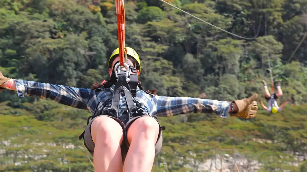 Two people ziplining above a forest. A woman is in the foreground and is sat upright, a man is in the background hanging upside down.