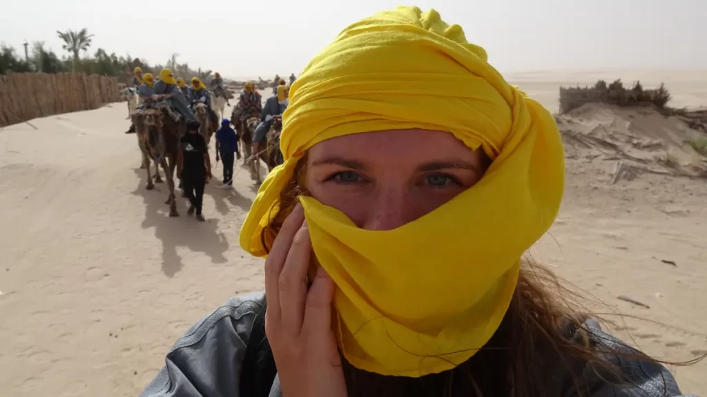 Woman in a yellow headscarf on a camel in a sandy desert with several other people behind her also on camels