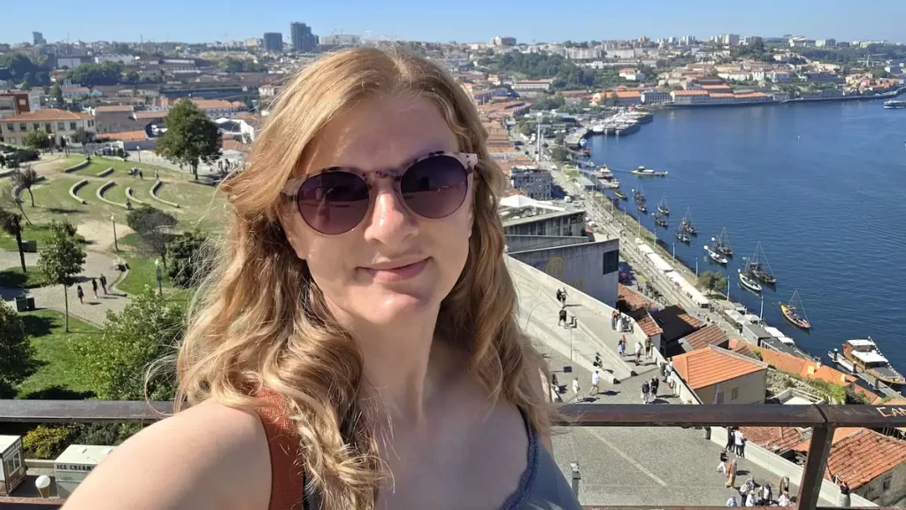 Blonde haired woman wearing sunglasses taking a selfie with a river behind her and the Porto skyline