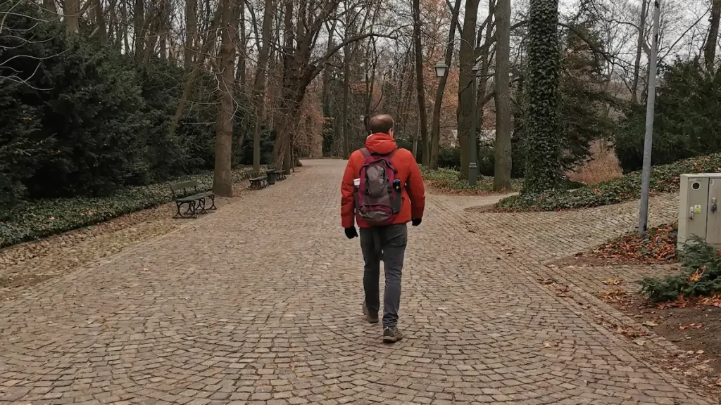 Man walking along a cobbled stone path in a park carrying a small backpack and wearing a red winter jacket and walking boots